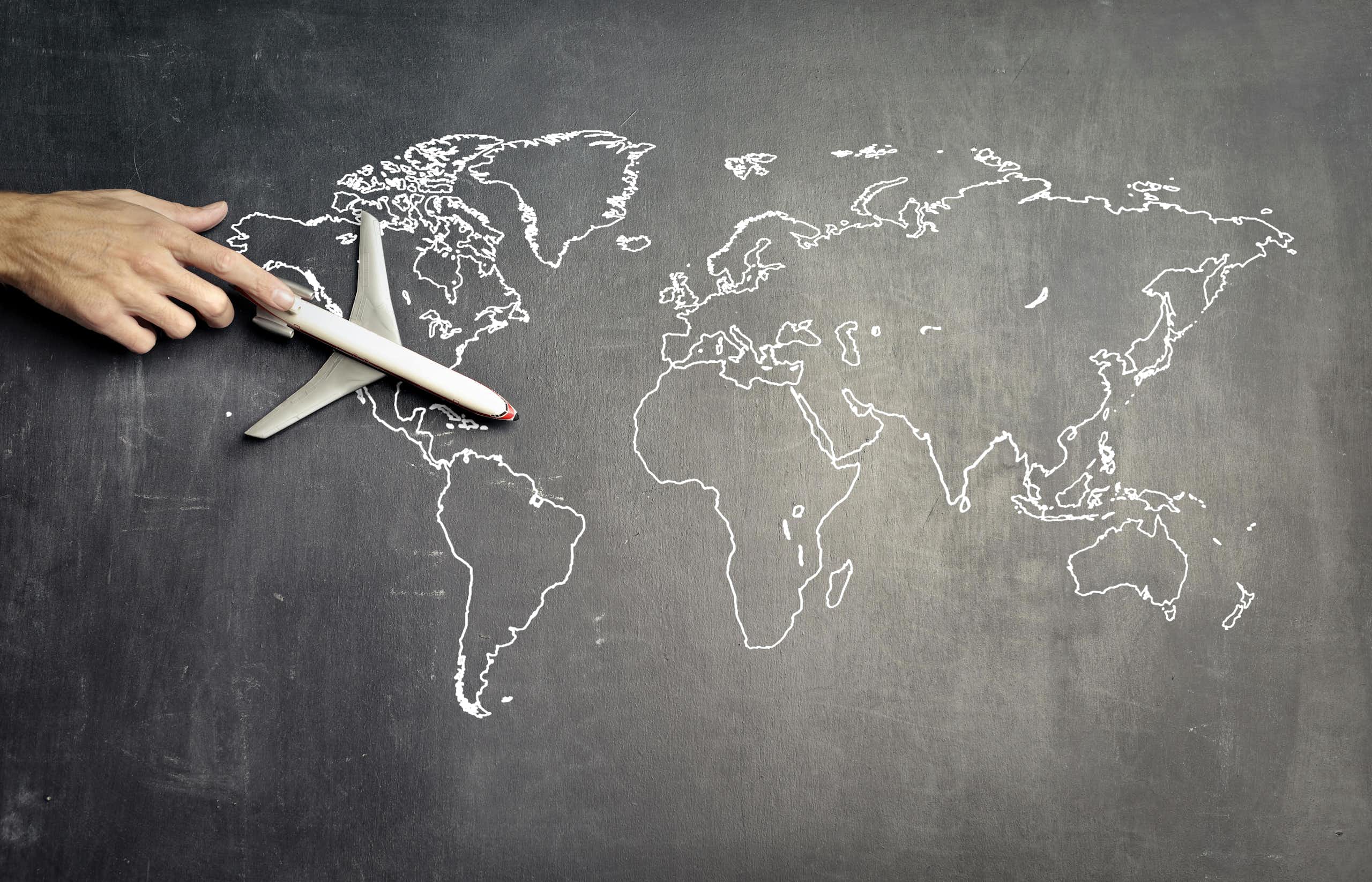 A hand pushes a toy plane over a world map on a blackboard