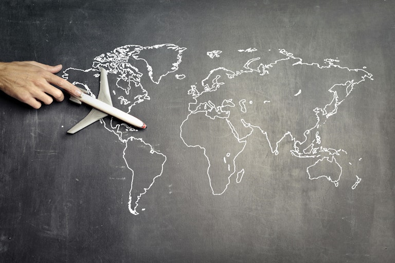 A hand pushes a toy plane over a world map on a blackboard
