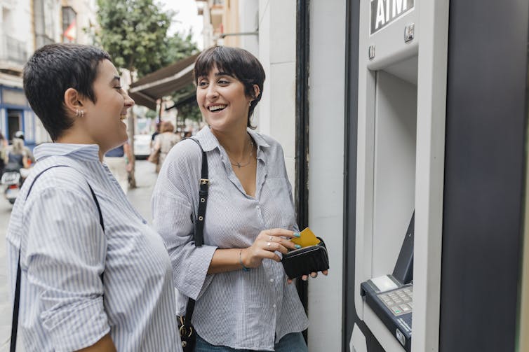 Two women in button-down shirts stand chatting and smiling next to an ATM on an outside wall.