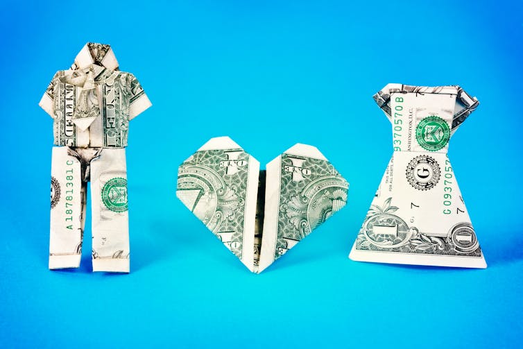 Two silhouettes made of dollar bills – one in pants, the other in a dress – stand on either side of a heart made of a dollar bill.
