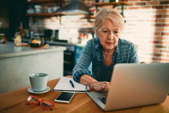 Older woman at laptop in kitchen, with cup/saucer, smartphone, pen and paper on table