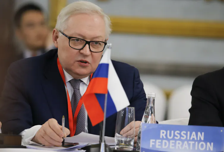A man with glasses and white hair sat at a desk behind a Russian flag.