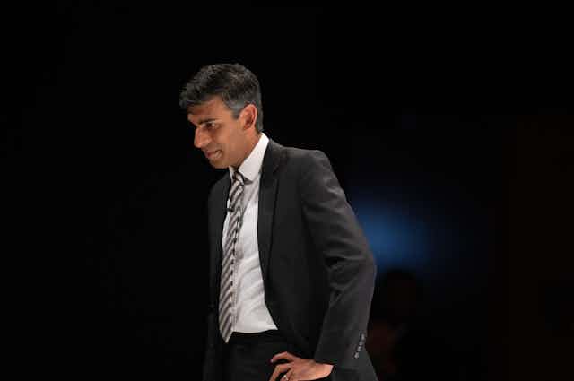Rishi Sunak in a suit against a dark background, looking down with his hand on his hip
