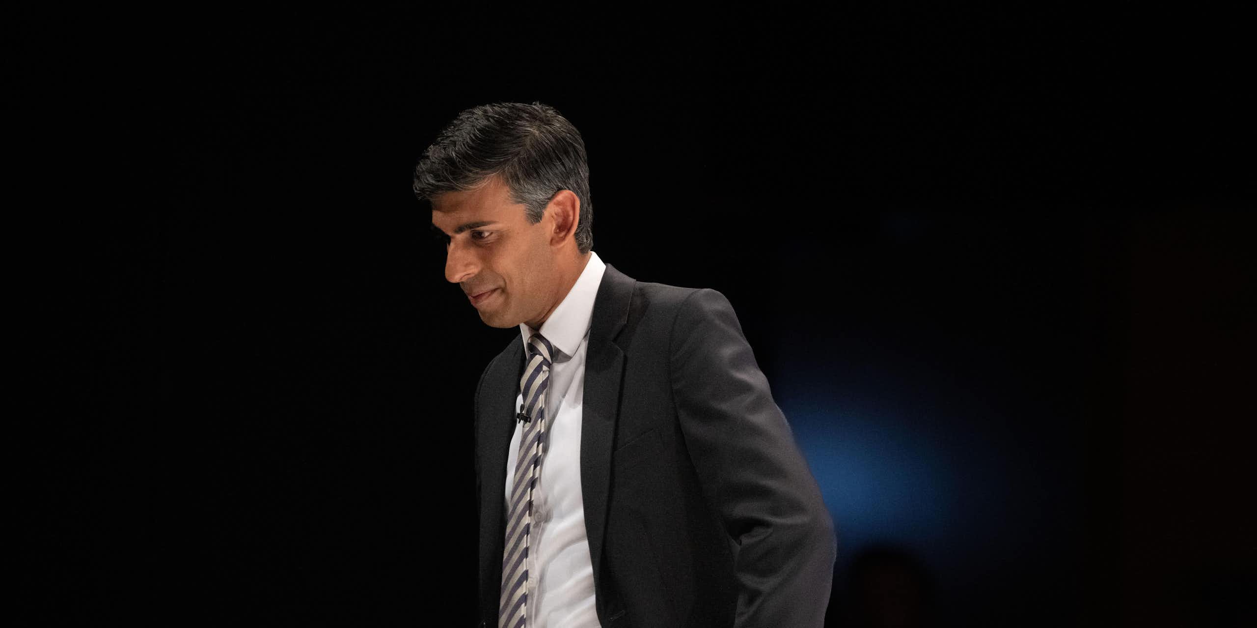 Rishi Sunak in a suit against a dark background, looking down with his hand on his hip