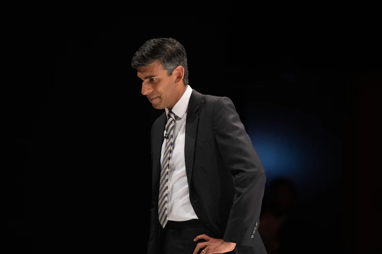 Rishi Sunak in a suit against a dark background, looking down with his hand on his hip