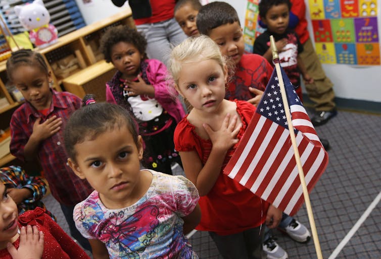 Children stand behind the American flag with one hand over their heart.