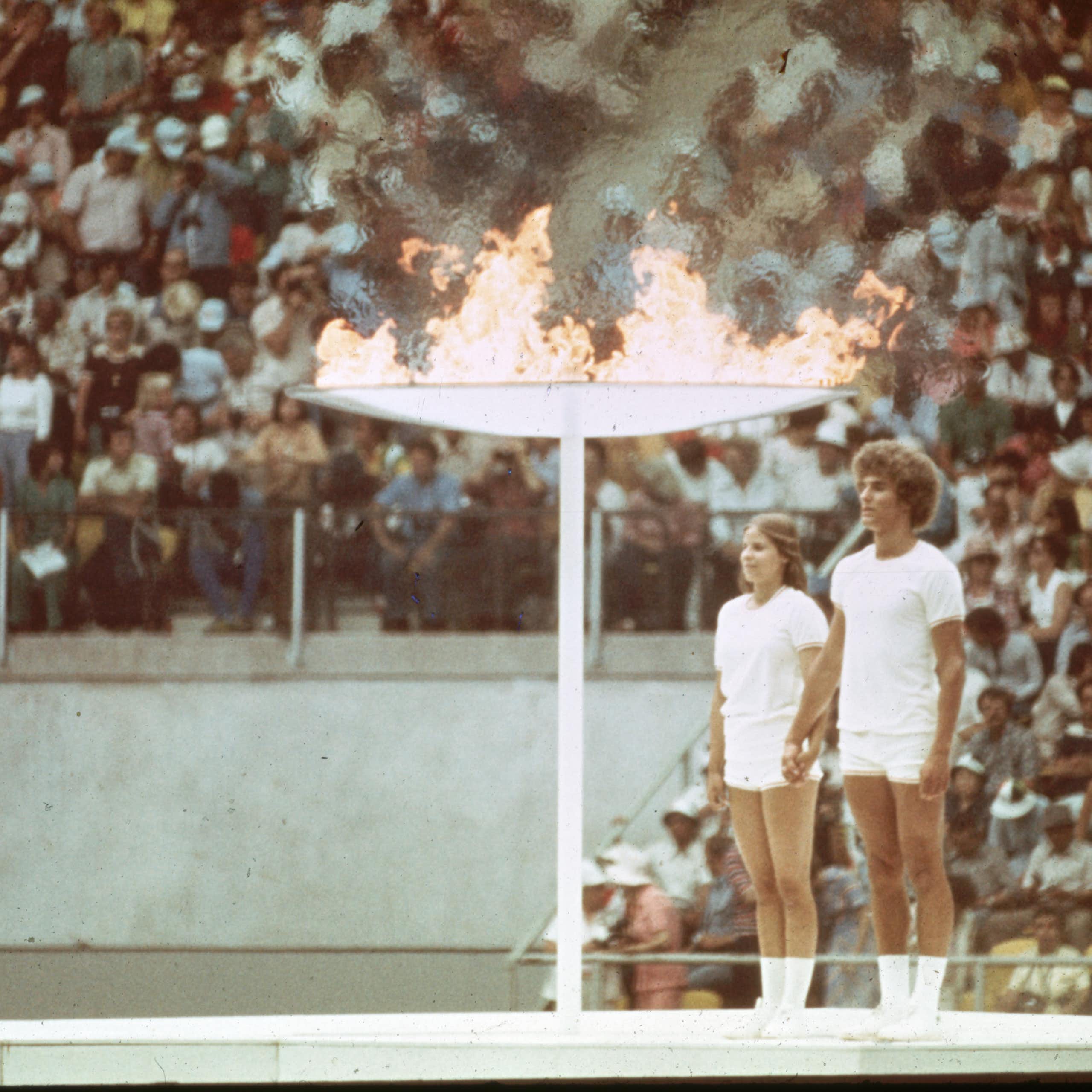A vintage colour photo of two young people - a man and woman - standing at attention next to a giant torch that is flaming. In the background, packed stands of a stadium.