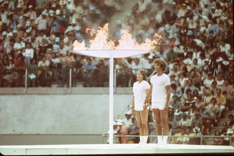 A vintage colour photo of two young people - a man and woman - standing at attention next to a giant torch that is flaming. In the background, packed stands of a stadium.