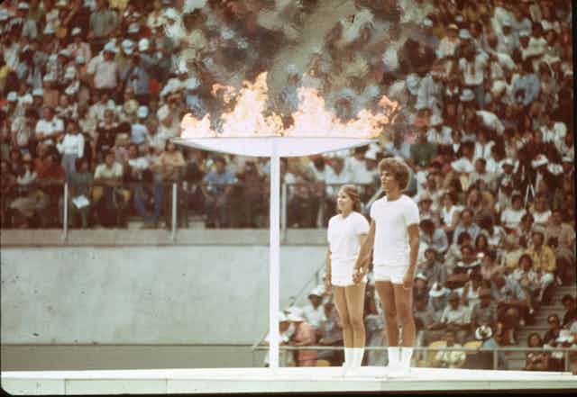 Photo couleur d'époque de deux jeunes gens - un homme et une femme - au garde-à-vous près d'une torche géante enflammée. A l'arrière-plan, les gradins d'un stade sont remplis.