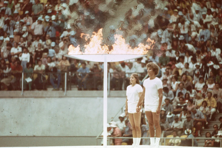 Photo couleur d'époque de deux jeunes gens - un homme et une femme - au garde-à-vous près d'une torche géante enflammée. A l'arrière-plan, les gradins d'un stade sont remplis.