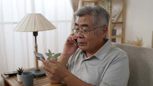 A man with gray hair and glasses sits inside on a couch, talking on a cell phone, while he looks at a credit card he is holding.
