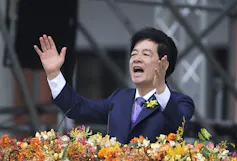 An Asian man in a blue suit and purple tie holds his hands up as he speaks behind a bank of brightly coloured flowers.