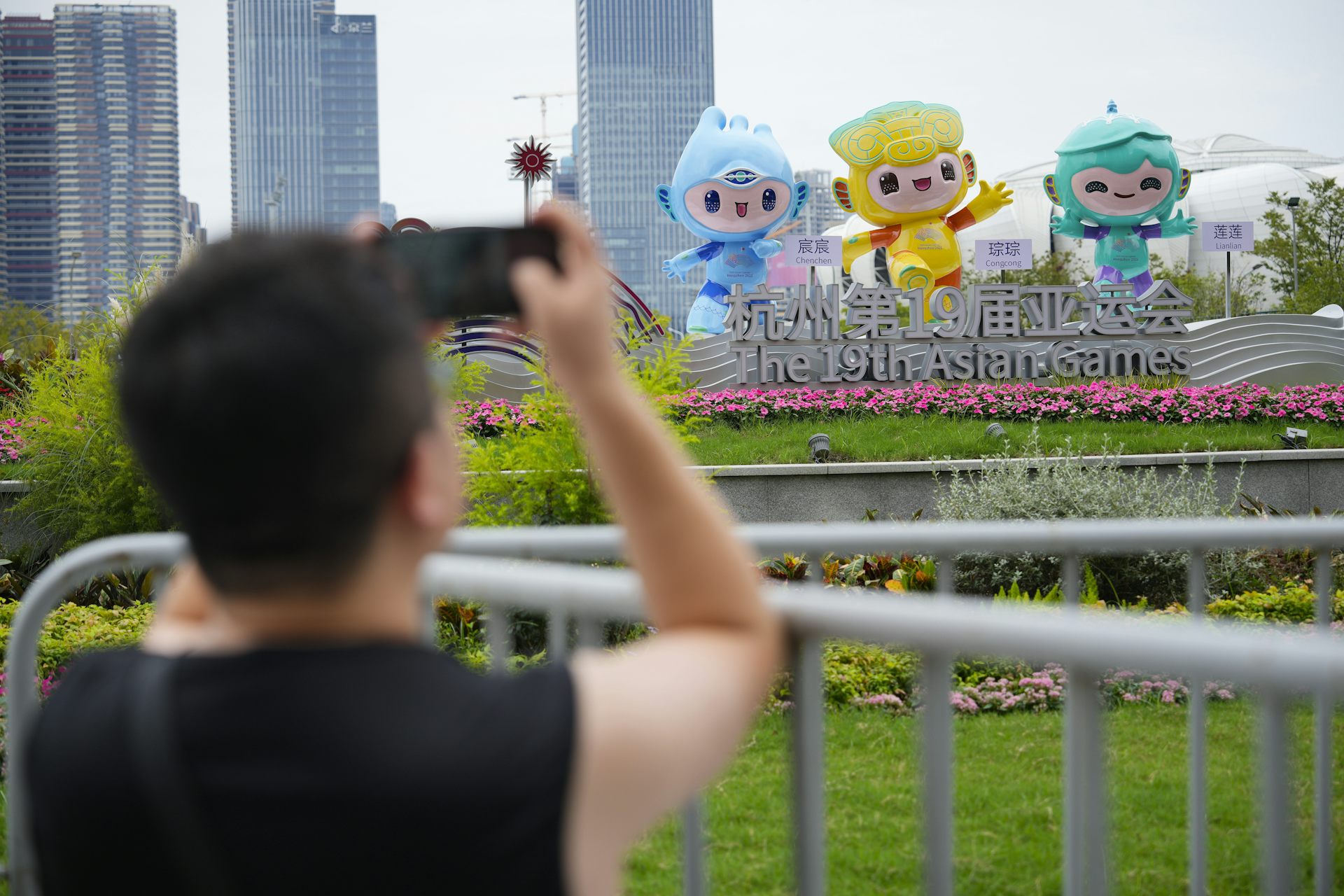 A man in a dark T-shirt takes photos of large colourful blow-up figures outside a stadium.