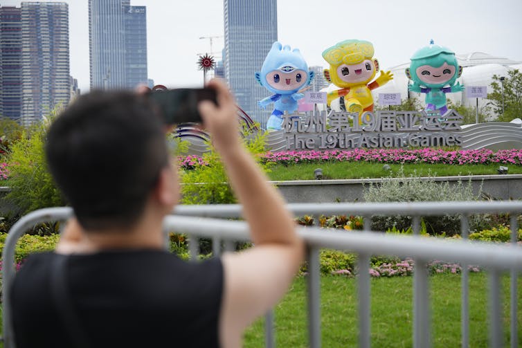 A man in a dark T-shirt takes photos of large colourful blow-up figures outside a stadium.