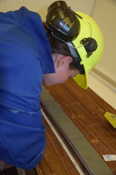A female scientist wearing a hard hat inspects mud layers in a sediment core.