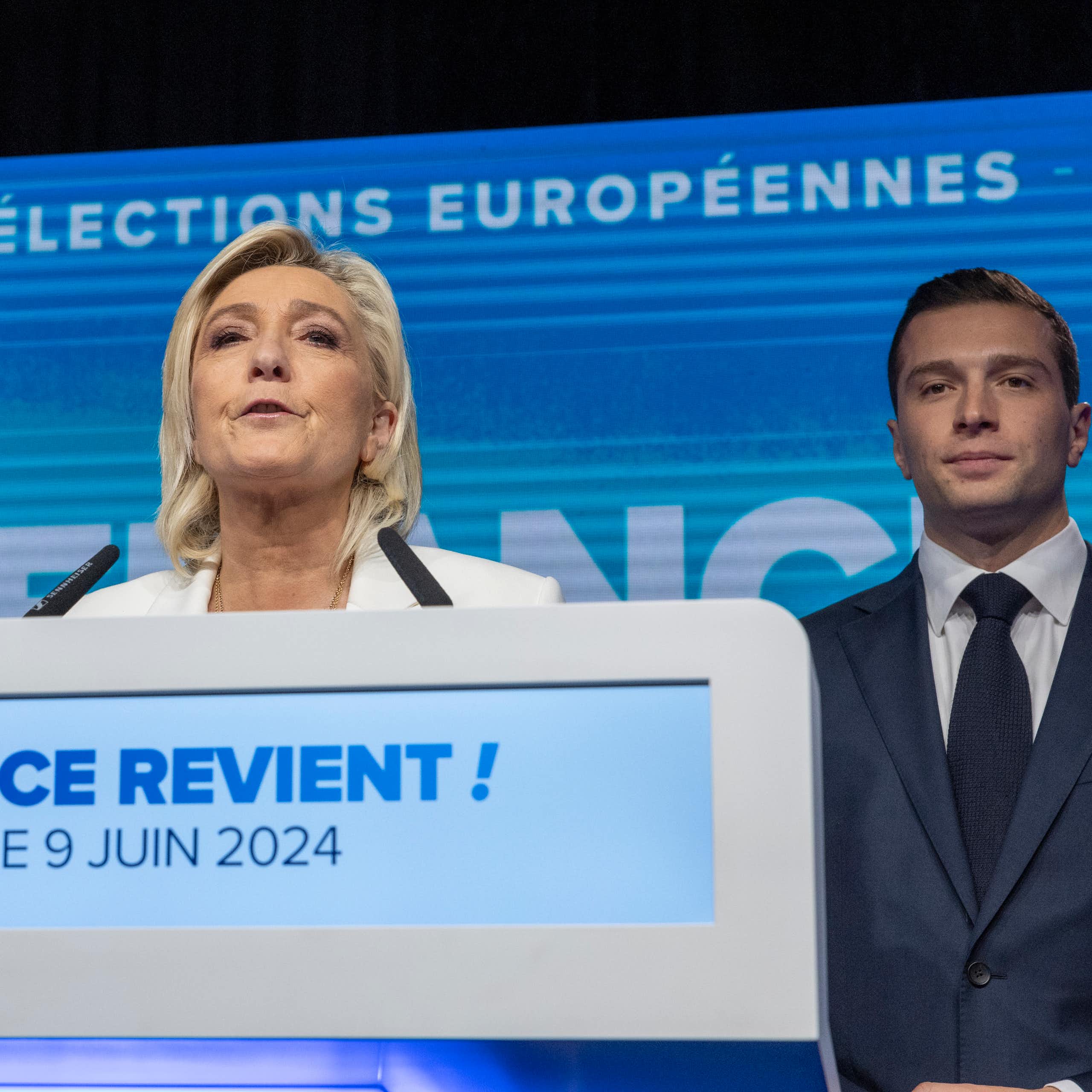 Marine Le Pen delivering a speech while a man in a suit stands in the background smiling.
