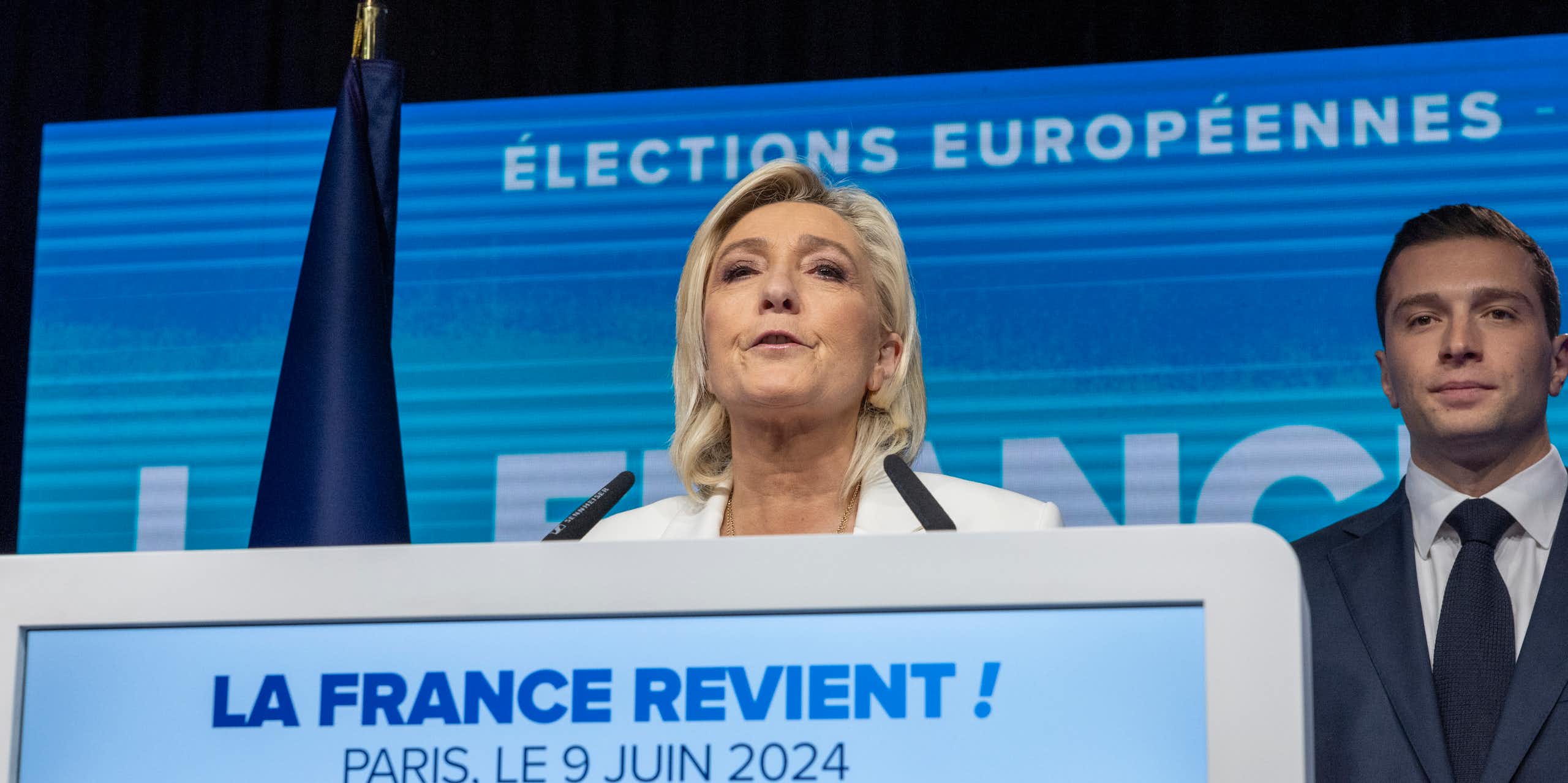 Marine Le Pen delivering a speech while a man in a suit stands in the background smiling.