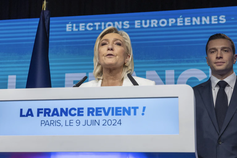 Marine Le Pen delivering a speech while a man in a suit stands in the background smiling.