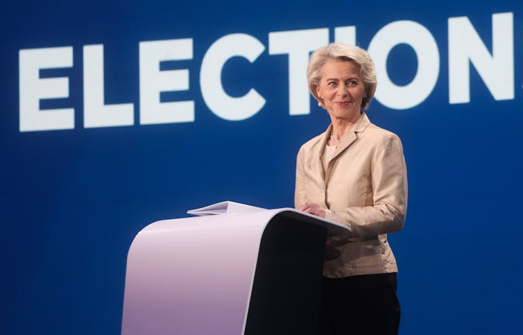 Von der Leyen standing at a podium in front of a sign reading