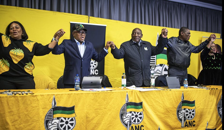 Three men and two women stand at a long table draped in yellow fabric with the ANC insignia. They are holding hands and appear to be singing or chanting.