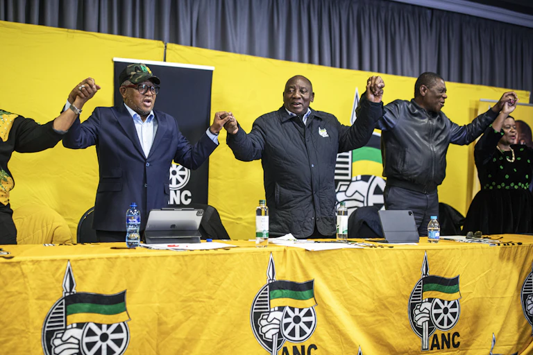 Three men and two women stand at a long table draped in yellow fabric with the ANC insignia. They are holding hands and appear to be singing or chanting.