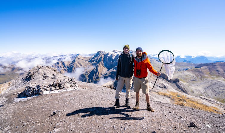 two men, one with big insect net in hand, on top of gravel mountain top, blue sky in background
