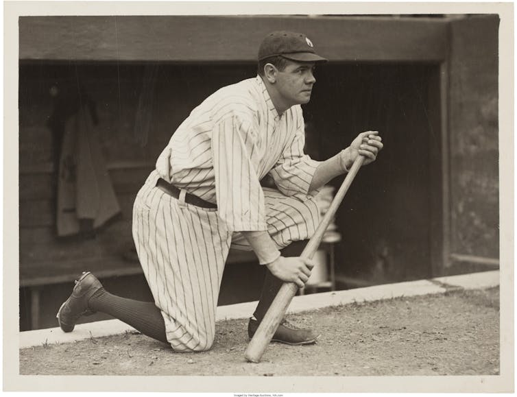 Babe ruth kneeling next to the dugout balancing on his bat.