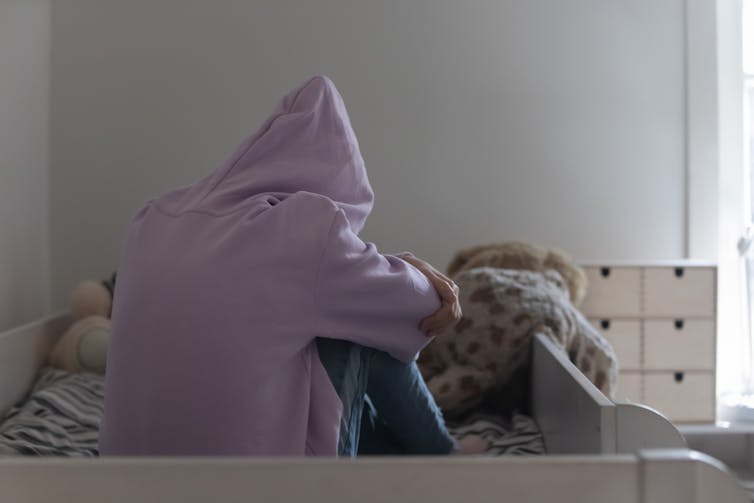 A lone teen wearing a lilac hoodie sits alone on a bed.