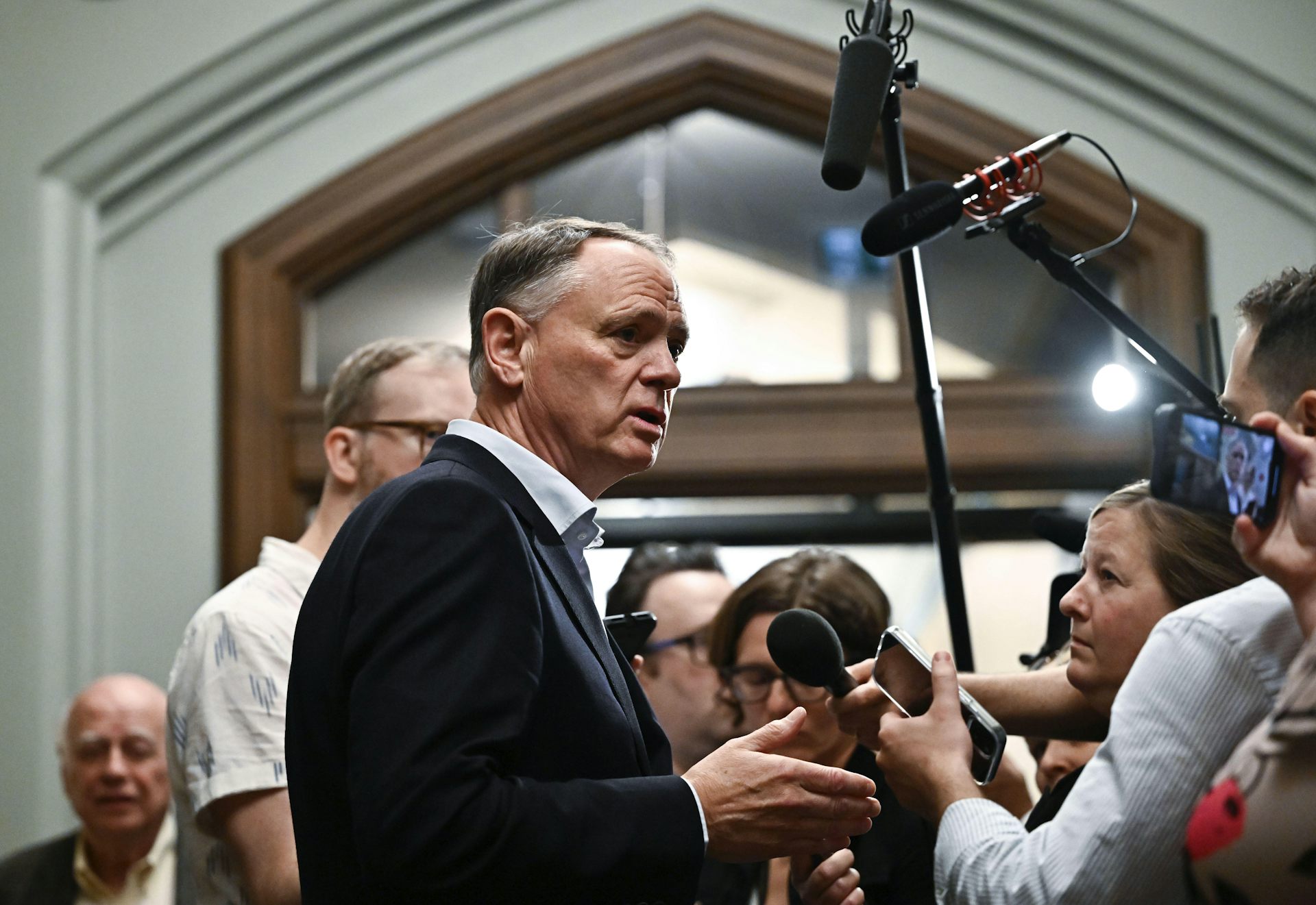 A man with thinning grey hair speaks amid a scrum of reporters, cameras and microphones.
