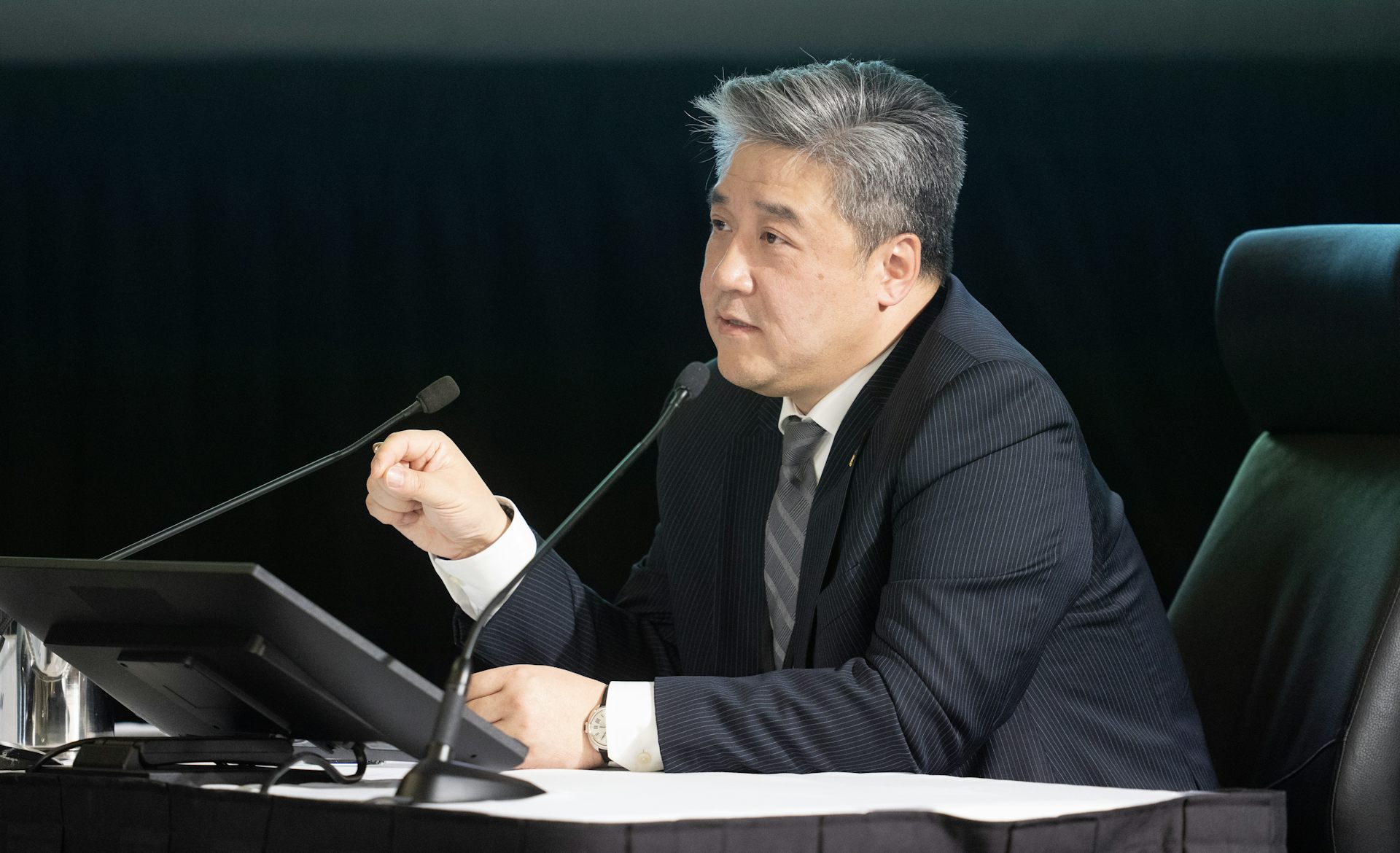 A man in a dark suit speaks into a microphone and gestures with his right hand.