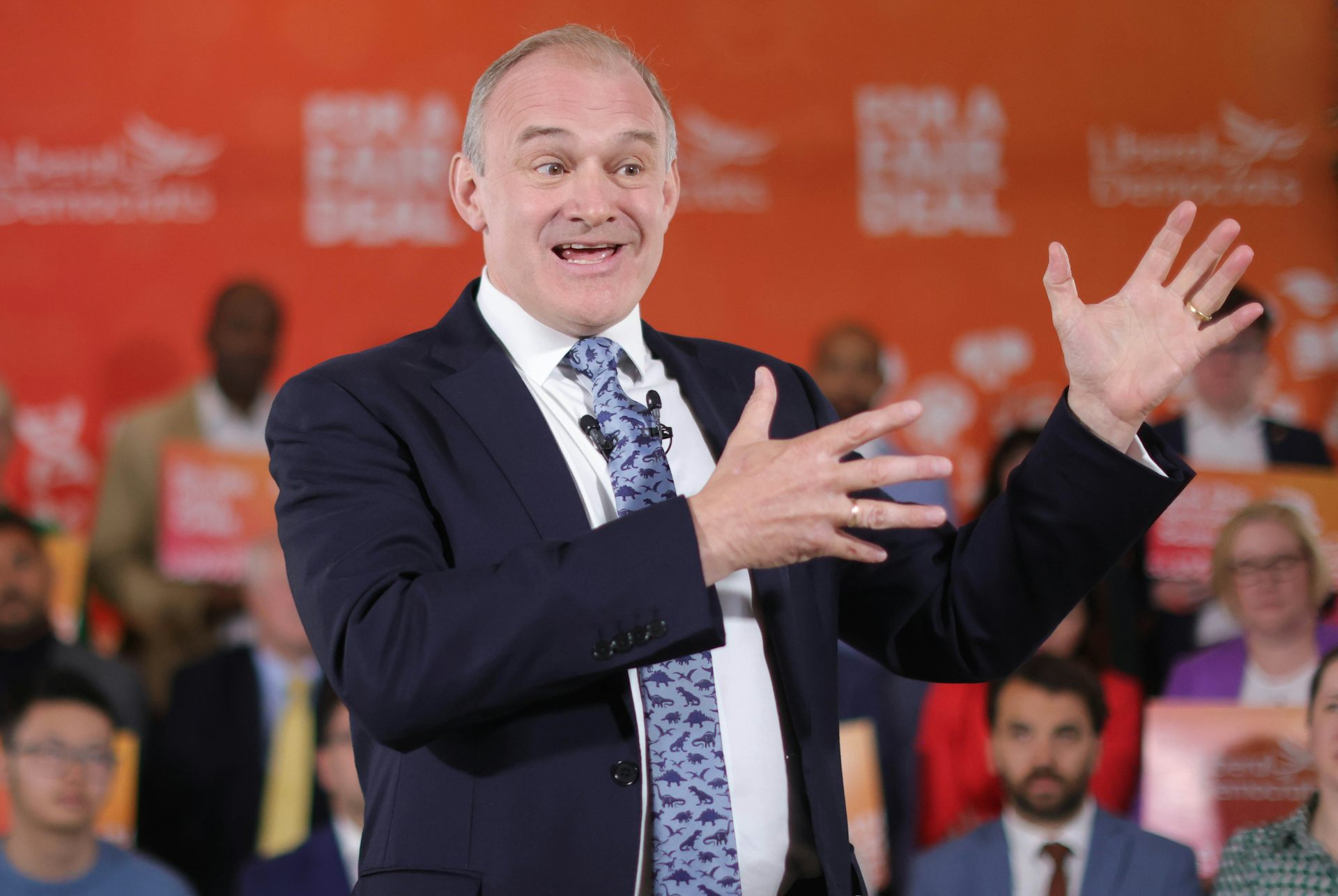 Ed Davey gesticulating in front of an orange background.