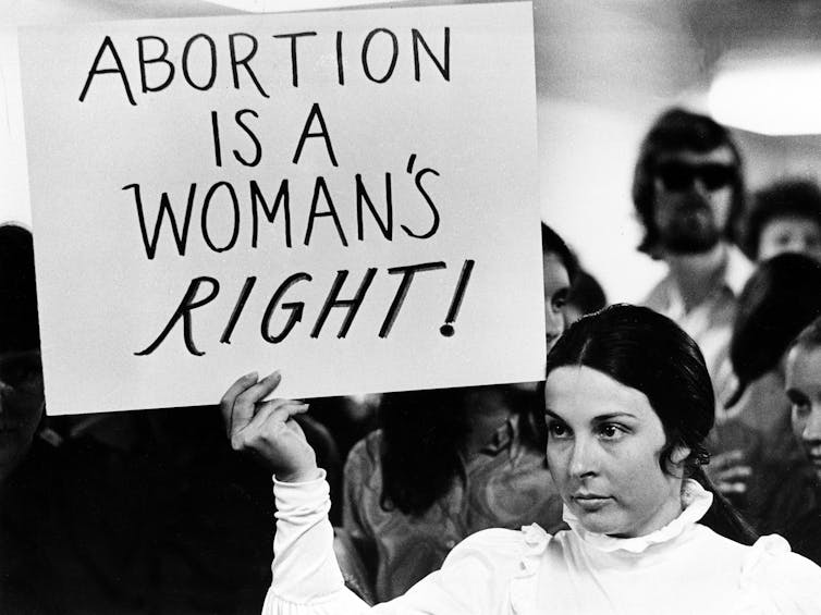 A black-and-white photograph shows a woman with dark hair and a white shirt holding a sign that reads, “Abortion is a woman’s right.”