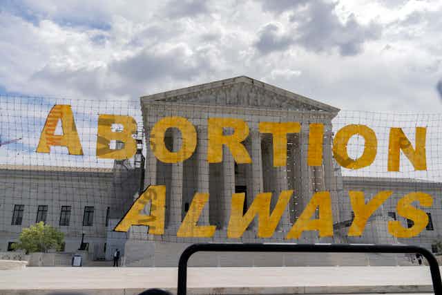 A large yellow sign says, 'Abortion always,' and is hung on a net in front of the Supreme Court building. 