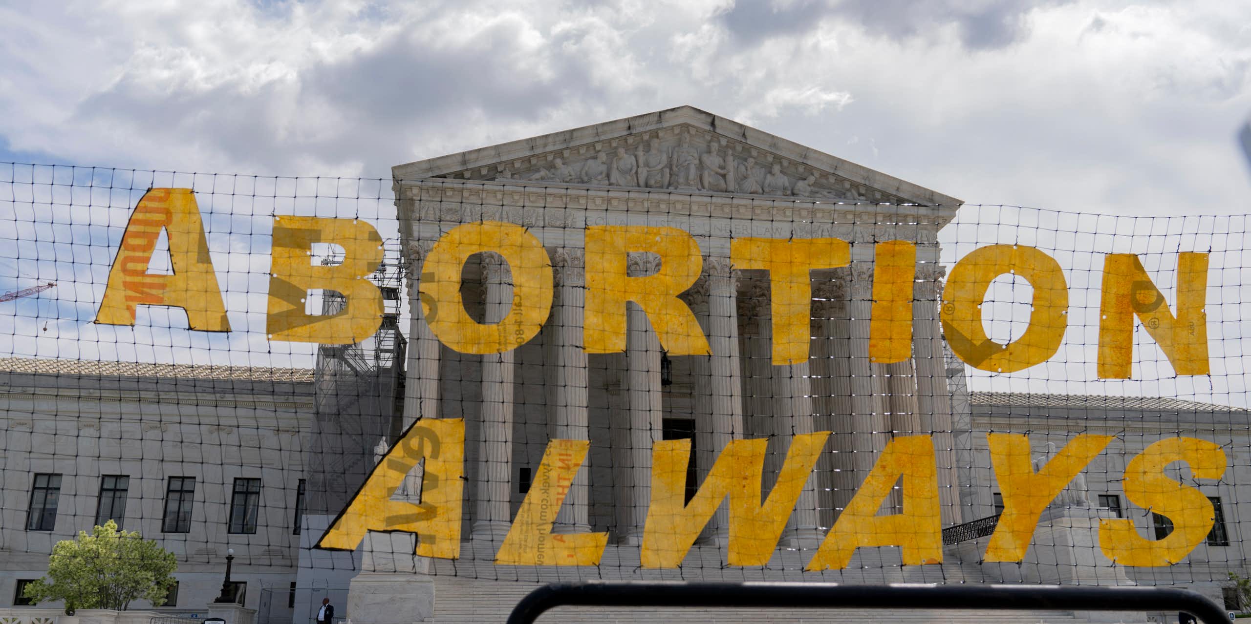 A large yellow sign says, 'Abortion always,' and is hung on a net in front of the Supreme Court building. 