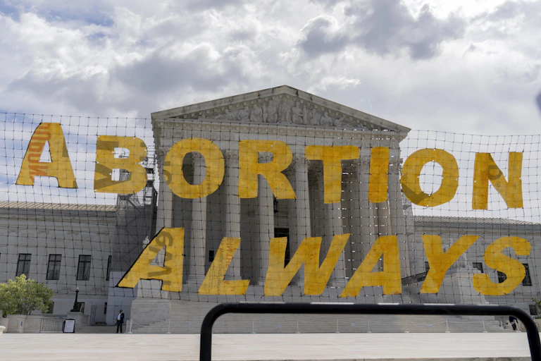 A large yellow sign says, 'Abortion always,' and is hung on a net in front of the Supreme Court building.