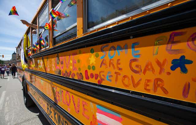 A school bus at a pride parade. People abord wave rainbow flags out the window. writing on the bus reads; some people are gay, get over it. 