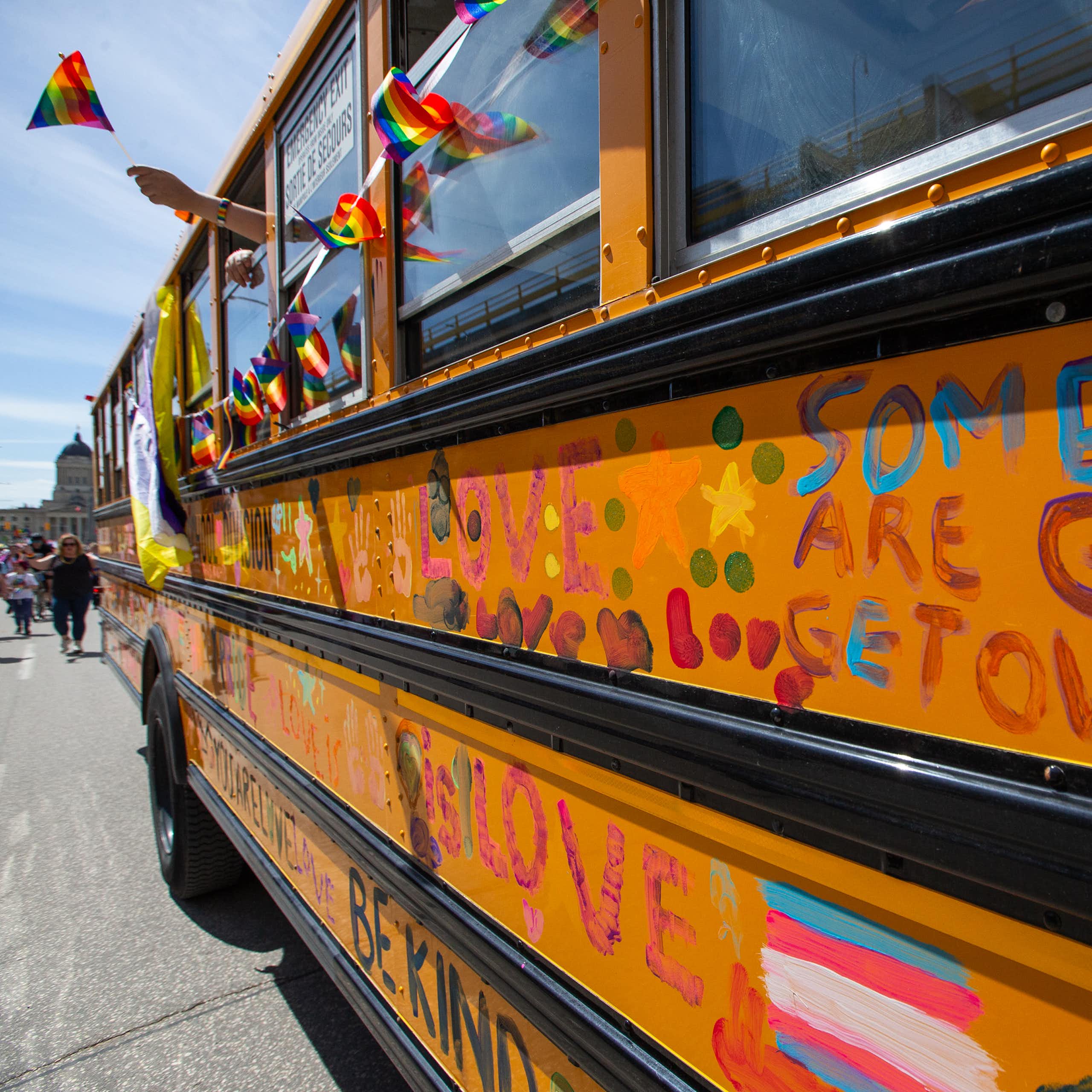 A school bus at a pride parade. People abord wave rainbow flags out the window. writing on the bus reads; some people are gay, get over it. 