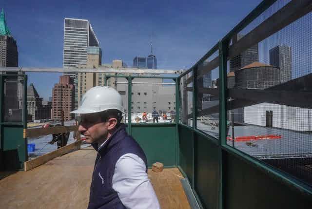 A man in a hard hat walks across a rooftop with skyscrapers behind him