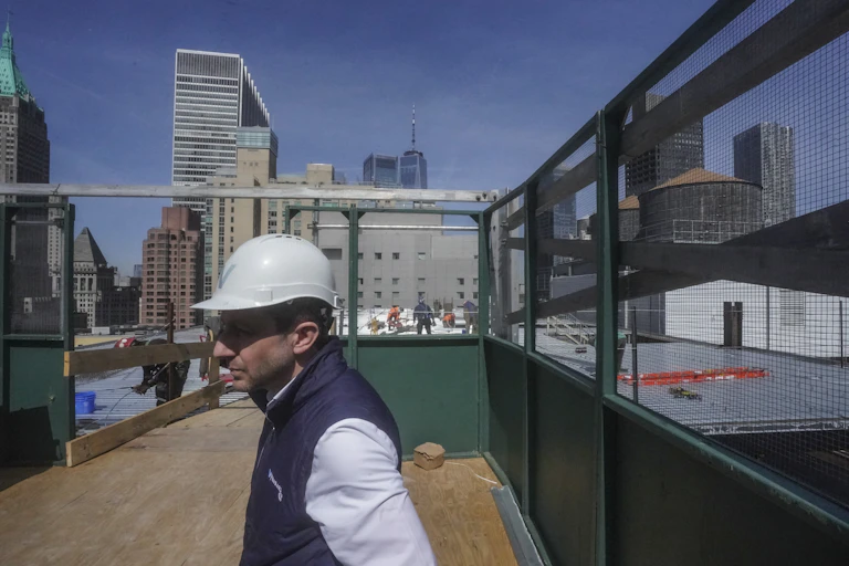 A man in a hard hat walks across a rooftop with skyscrapers behind him