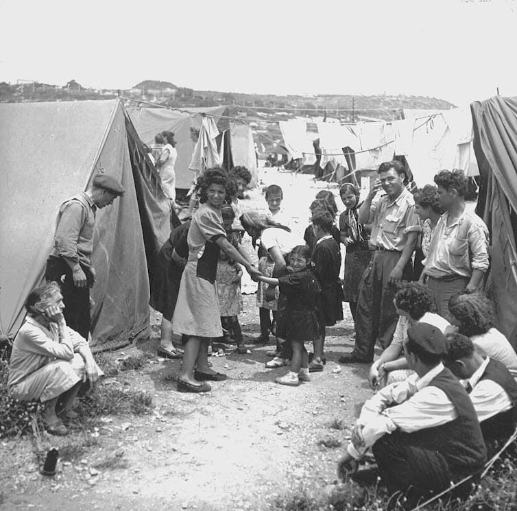 A black and white photo of around a dozen people sitting and standing between simple tents outside.