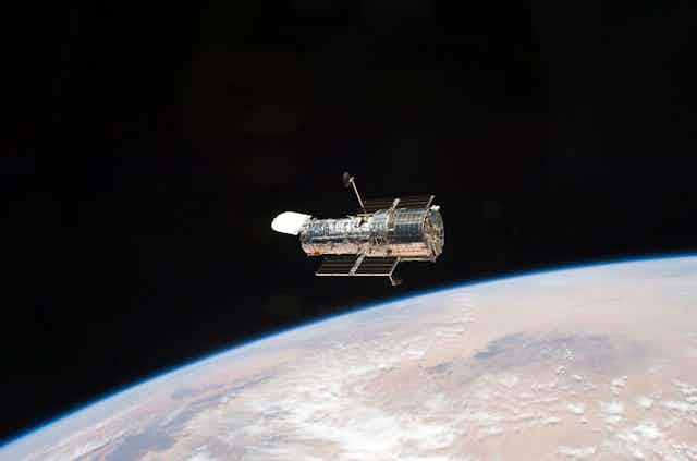 The Hubble telescope, which looks like a metal cylinder with solar panels attached, floating above Earth.