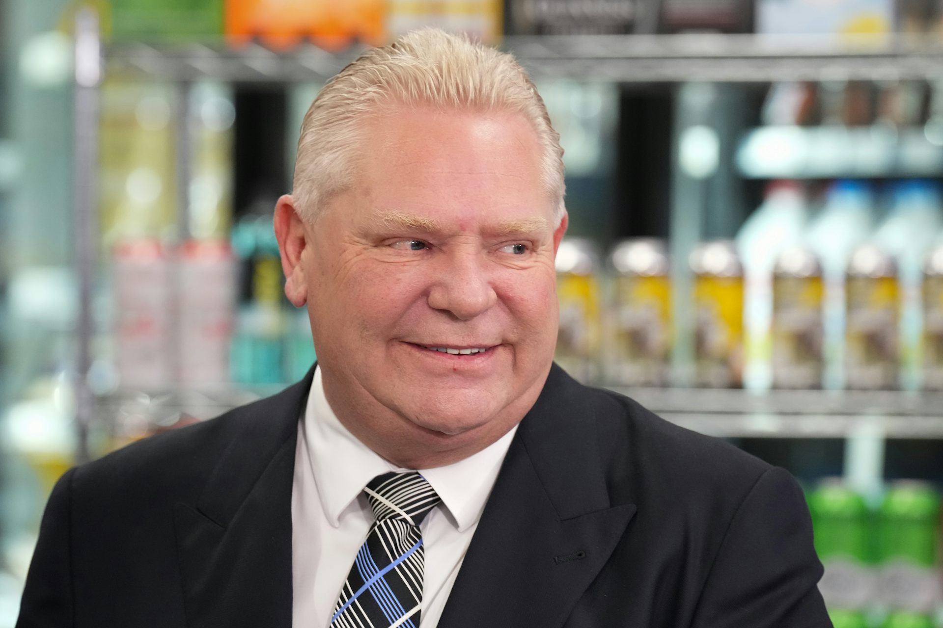 An older middle-aged man in a suit smiling while standing in front of a shelf full of canned alcoholic beverages