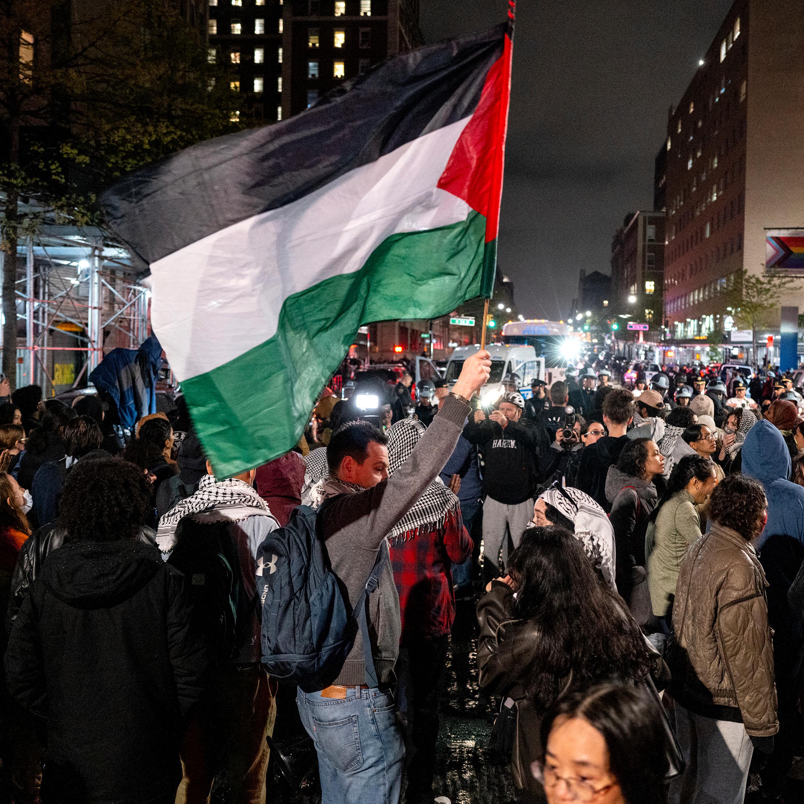 People gather in a city, with one of them holding a large Palestinian flag.