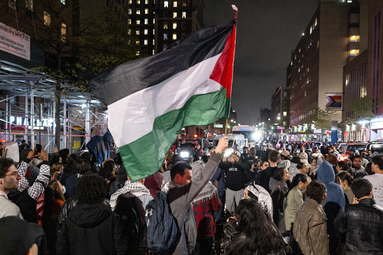 People gather in a city, with one of them holding a large Palestinian flag.