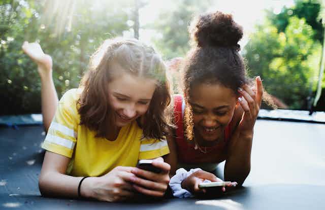 Two young, happy teen girls lie on a trampoline in a backyard looking at smartphones.