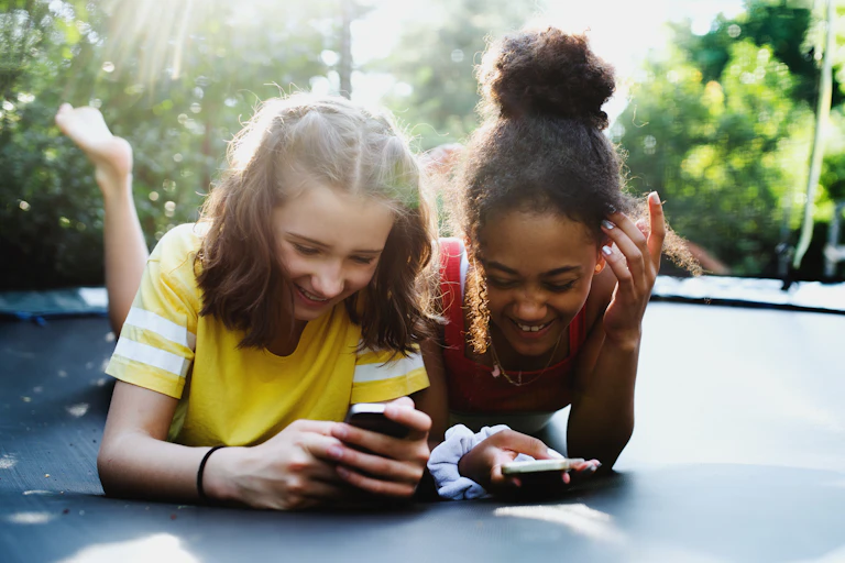 Two young, happy teen girls lie on a trampoline in a backyard looking at smartphones.