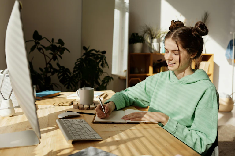 A teenager sits at a desk, writing in a notebook. In front is a large computer screen and keyboard.