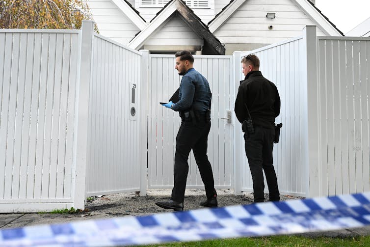 Two detectives stand outside a tall white gate surrounded by police cordon robbery cordon.