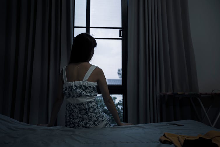 A clothed woman sits off camera on the edge of a bed in a darkened room