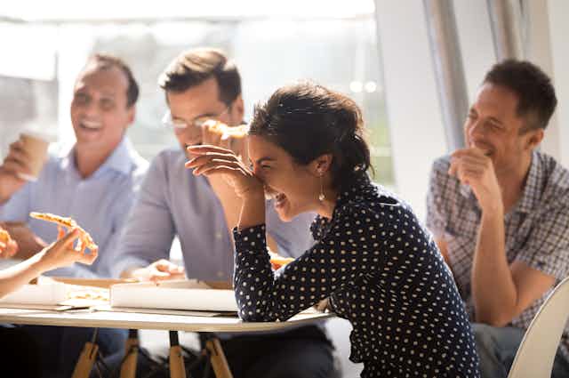 Woman and coworkers laugh at joke sitting around table with pizza
